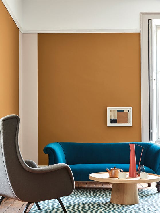 Living space painted in dark orange (Middle Buff) with a contrasting deep blue sofa, grey armchair and wooden table on a rug.