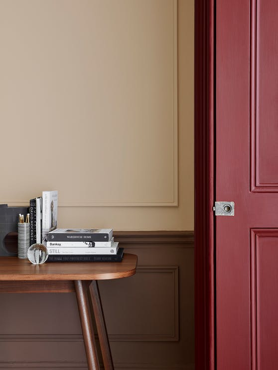 Close-up of a warm neutral paneled wall in Catel Pink, alongside a deep red door painted in Arras and a wooden desk.