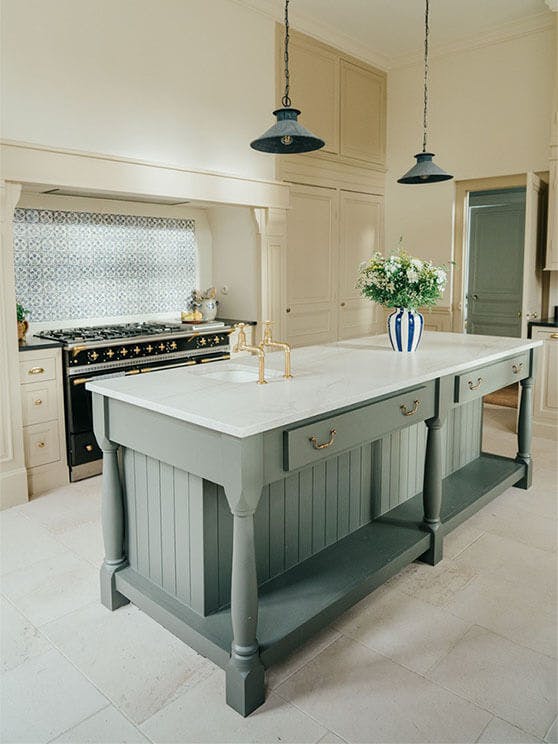 A large kitchen featuring a grey island in the middle with marble worktops, and walls painted in various neutral Clay tones.