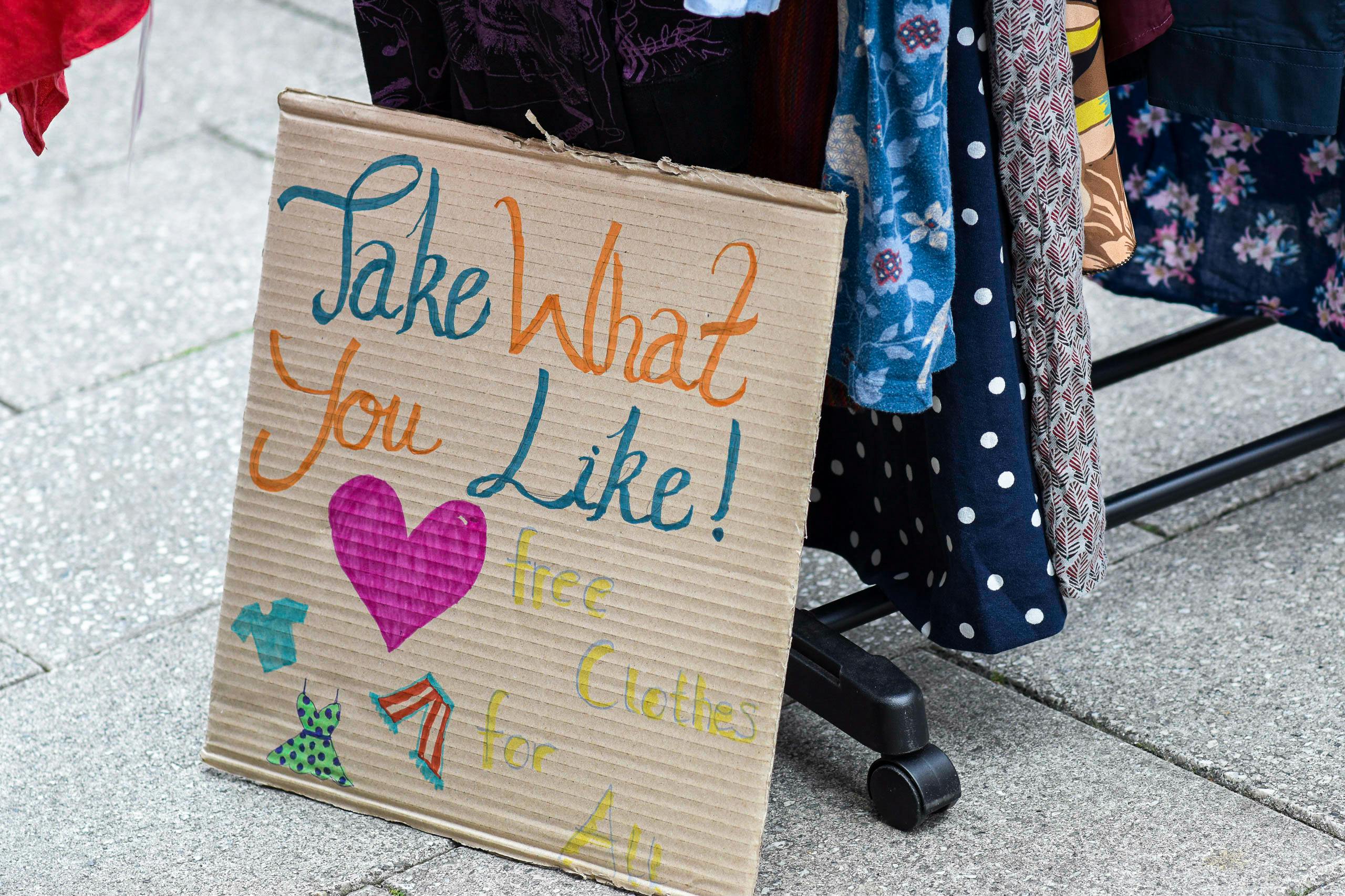 A sign saying "take what you like - free clothes for all" on the street next to a rack of free clothes.