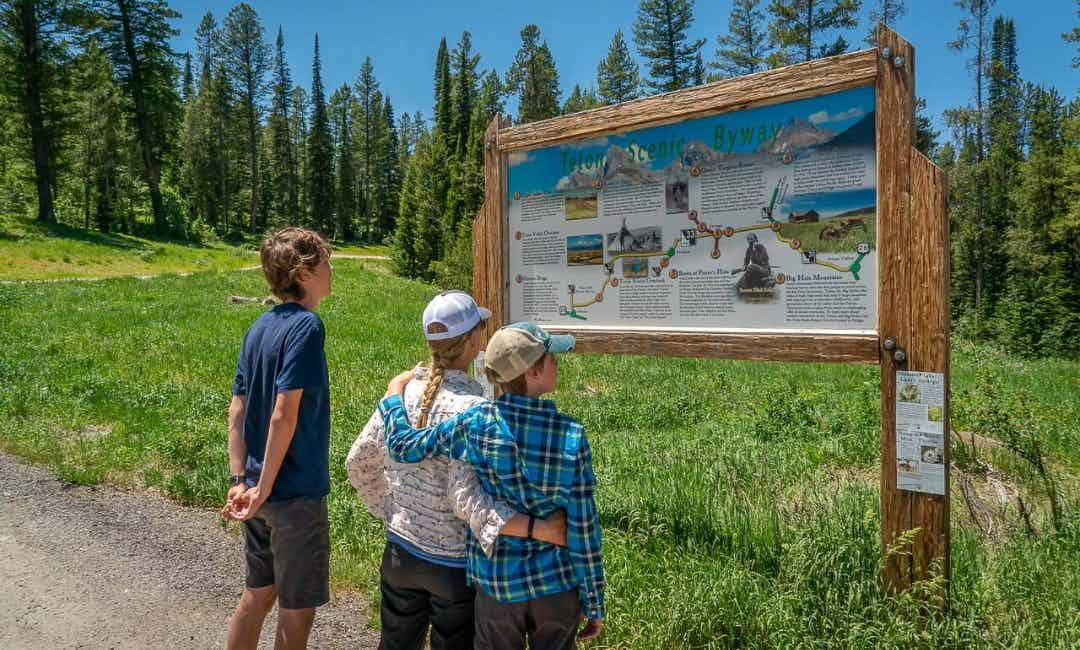 A family stands at the marker of the Teton Scenic Byway.