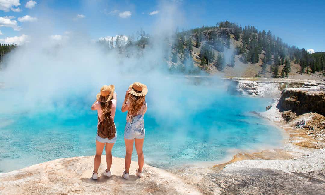 Two girls stand in front of the Great Prismatic Pond in Yellowstone National Park, a part of Yellowstone Teton Territory.