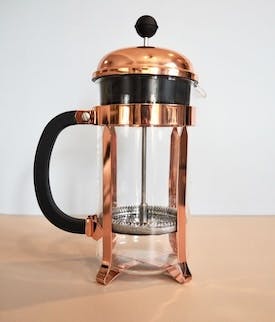 A French Press coffee brewer. The brewer is made from glass and has a metal frame with a handle for pouring. The lid has a plunger attached to it with a metal filter at the end.