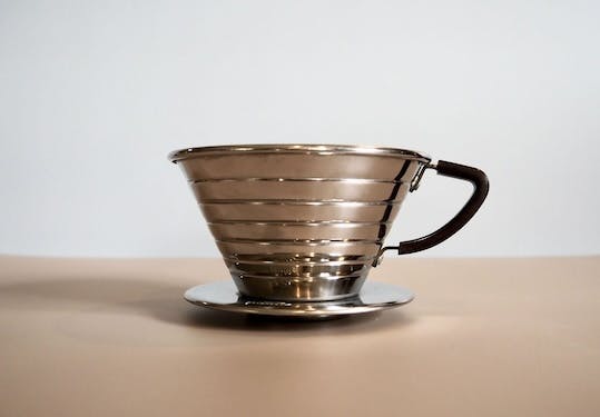 A Kalita Wave pour-over coffee brewer. It is made out of stainless steel and has a flat bottom with three holes for coffee to pass through. It has a handle.