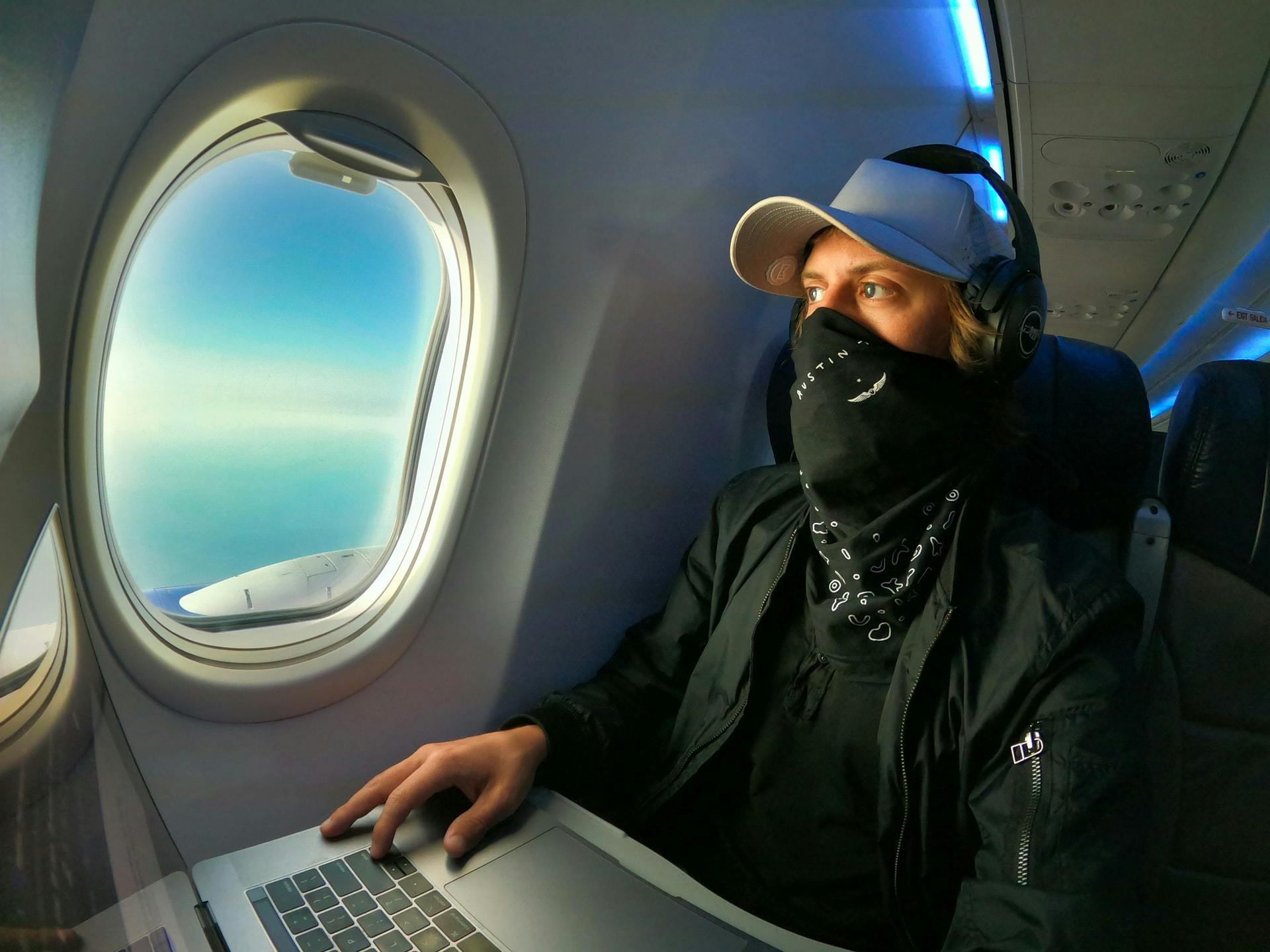 A man wearing a mask looking out of an airplane window and working on a laptop