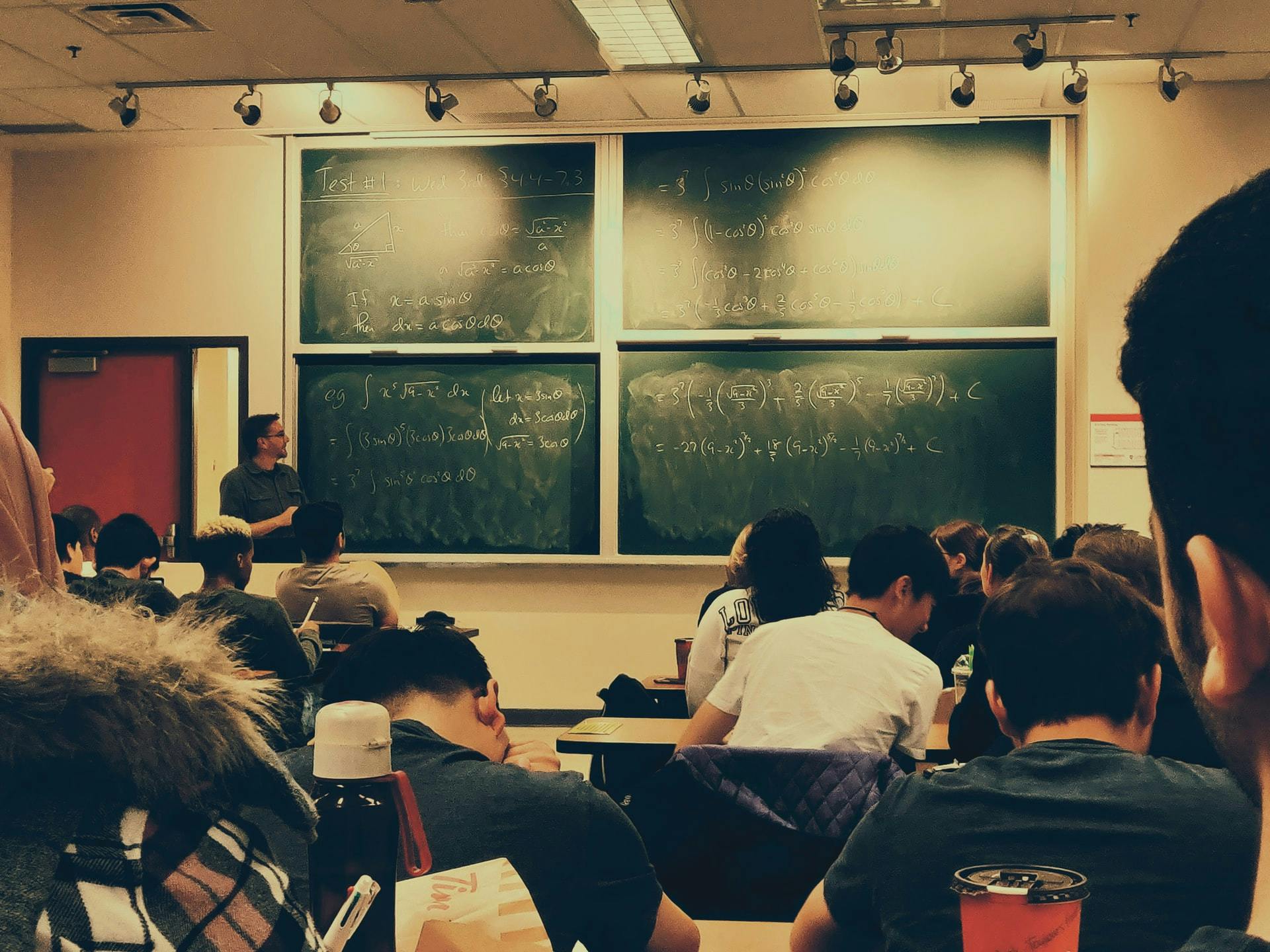 A teacher stands next to a blackboard and in front of a class of students