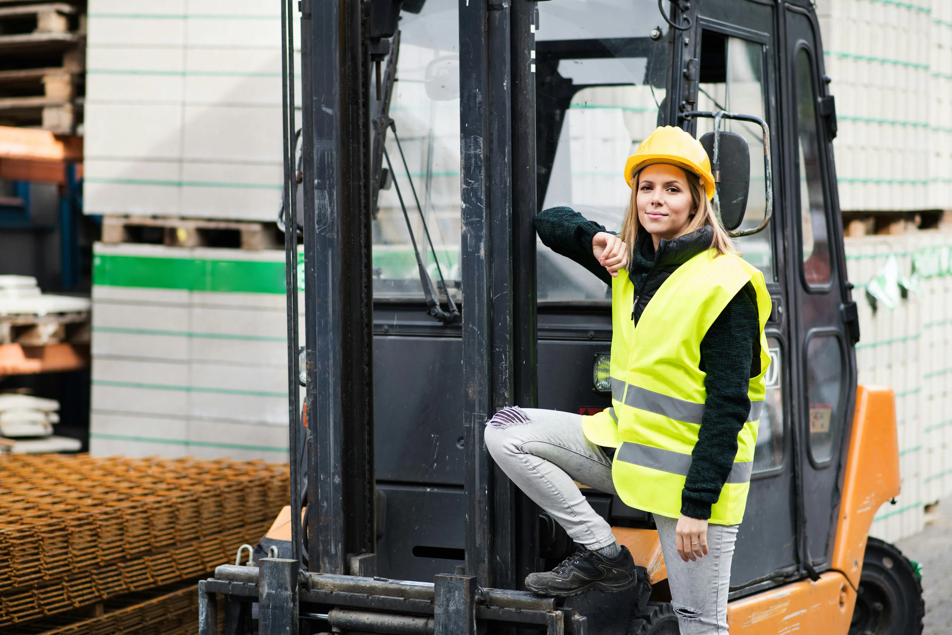 Warehouse worker in a hard hat and hi-vis vest standing at a forklift.
