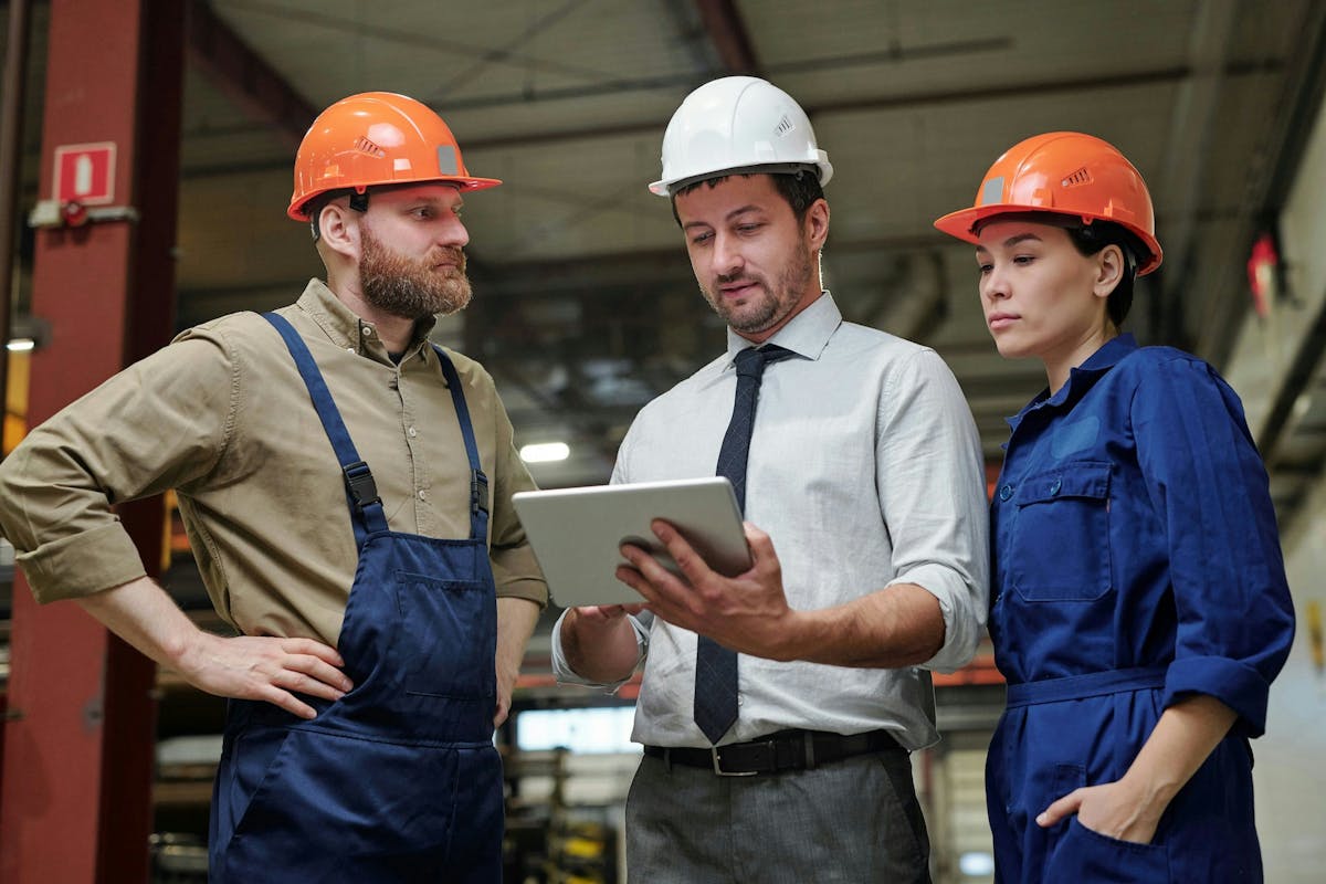 Factory supervisor in a white hard hat showing information on a tablet to two workers in orange helmets and blue overalls inside an industrial facility.