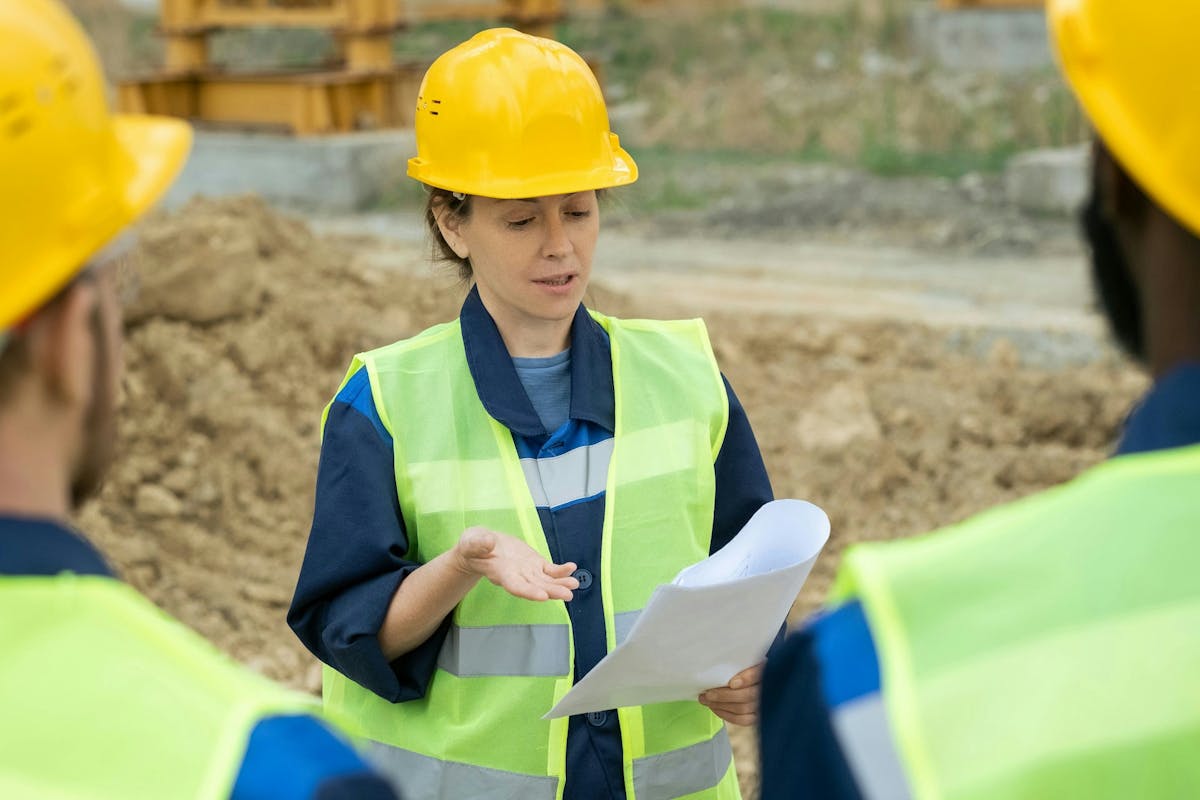 Construction supervisor in a yellow hard hat and safety vest holding documents and briefing workers at a construction site.