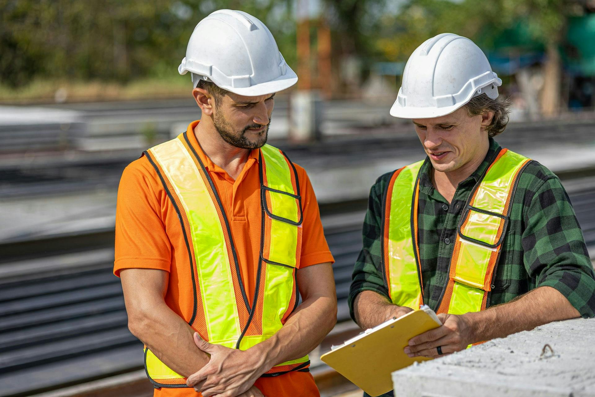 Two construction workers in white hard hats and reflective safety vests reviewing notes on a clipboard at an outdoor worksite.