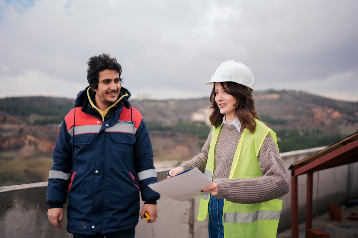 Two construction workers talking at a job site, one wearing a hard hat and reflective vest while holding documents, and the other in a blue jacket with red accents, standing outdoors with hills in the background.