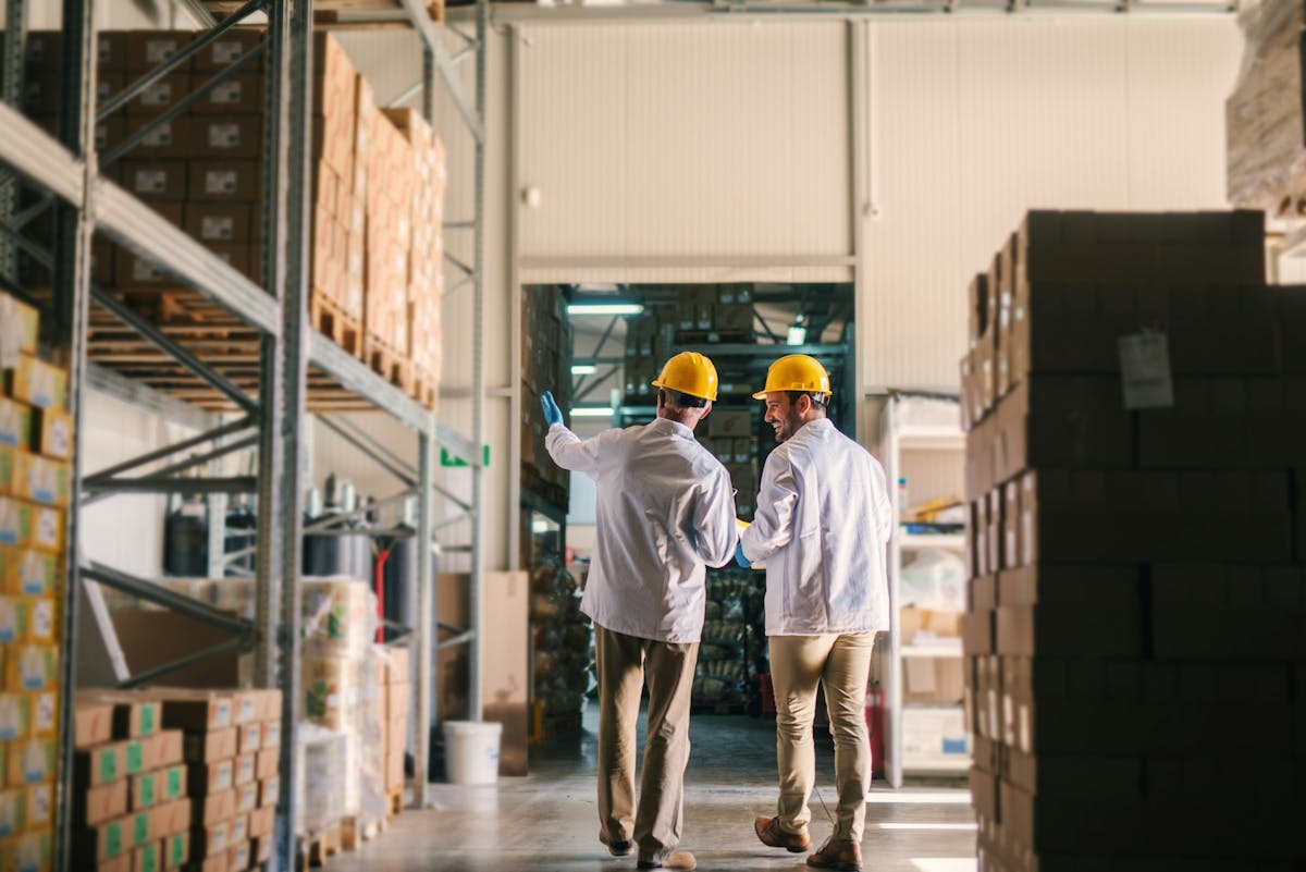 Two warehouse workers wearing hard hats and protective coats walk through a storage aisle