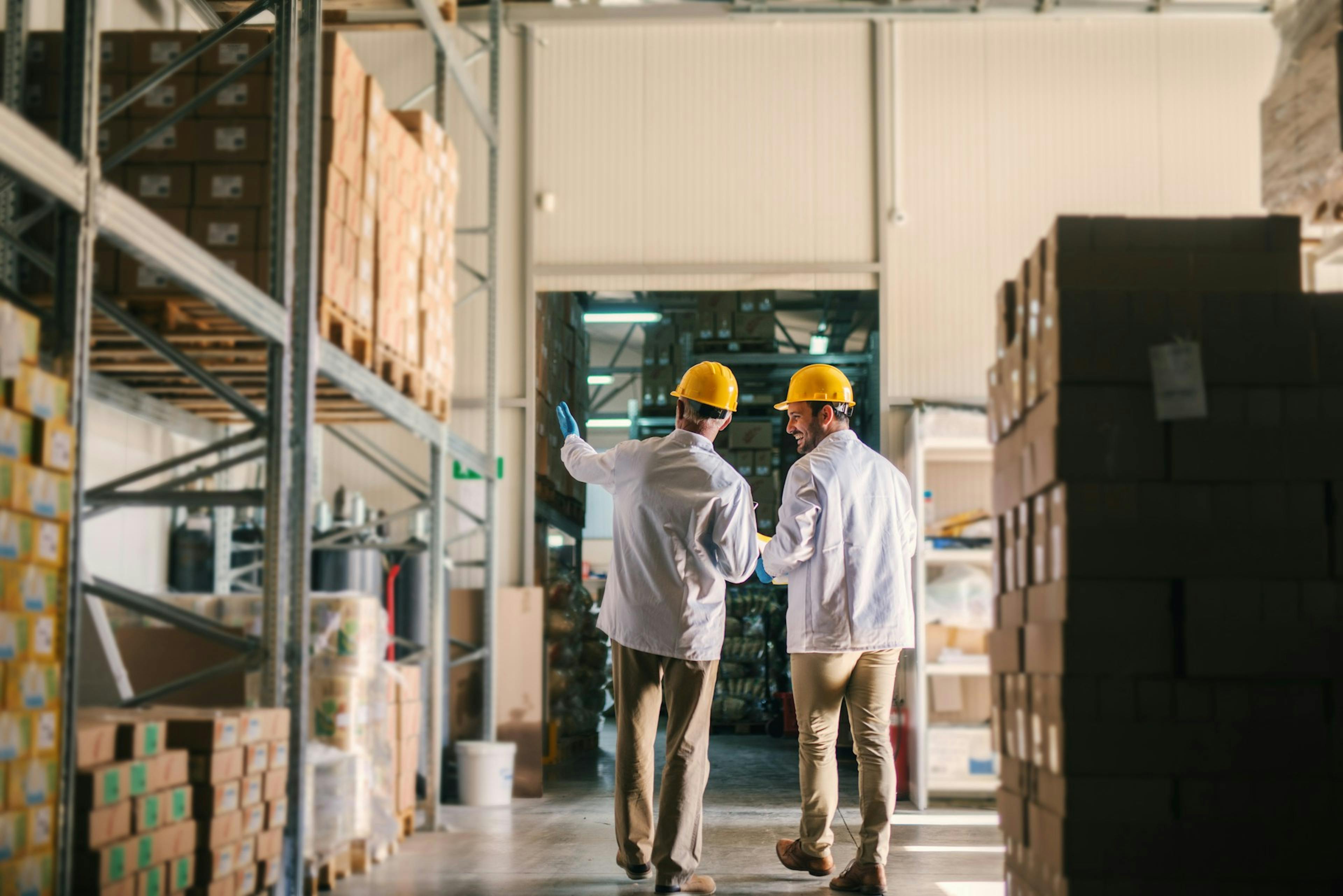Two warehouse workers wearing hard hats and protective coats walk through a storage aisle