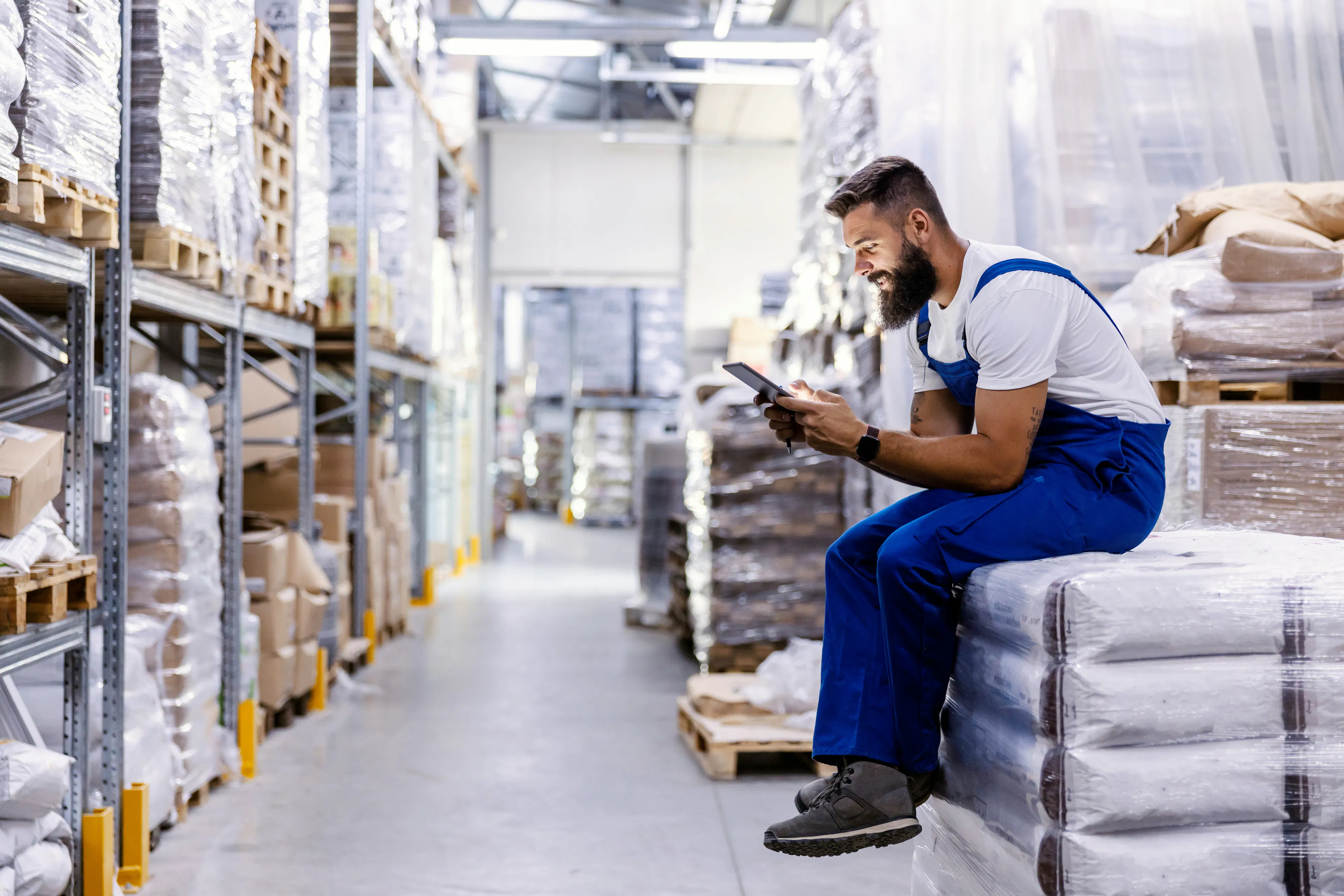 Warehouse worker checking handheld device while sitting on pallets