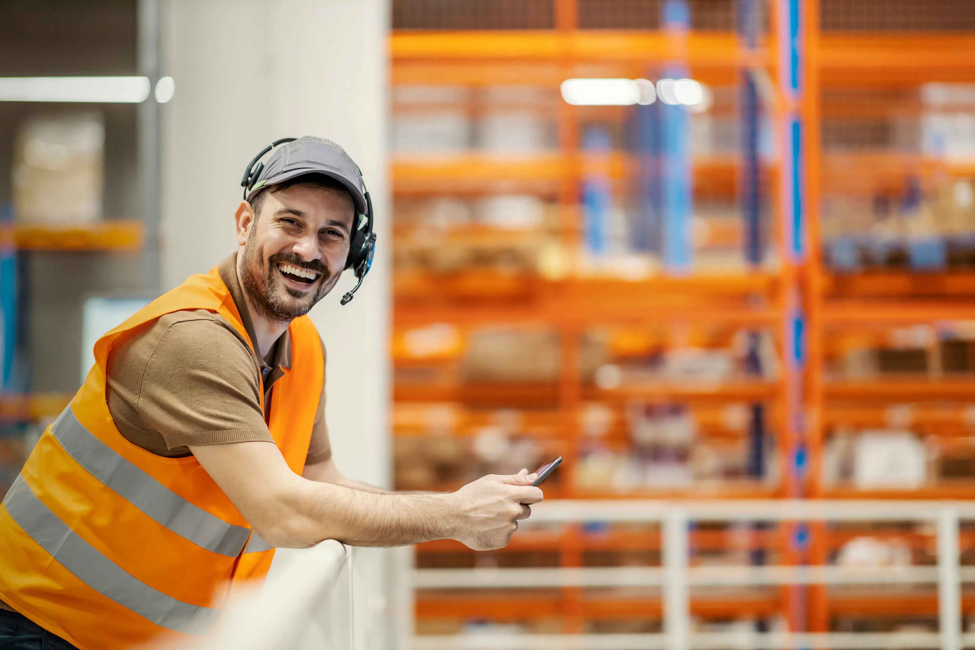 Smiling warehouse worker in an orange vest holding a phone
