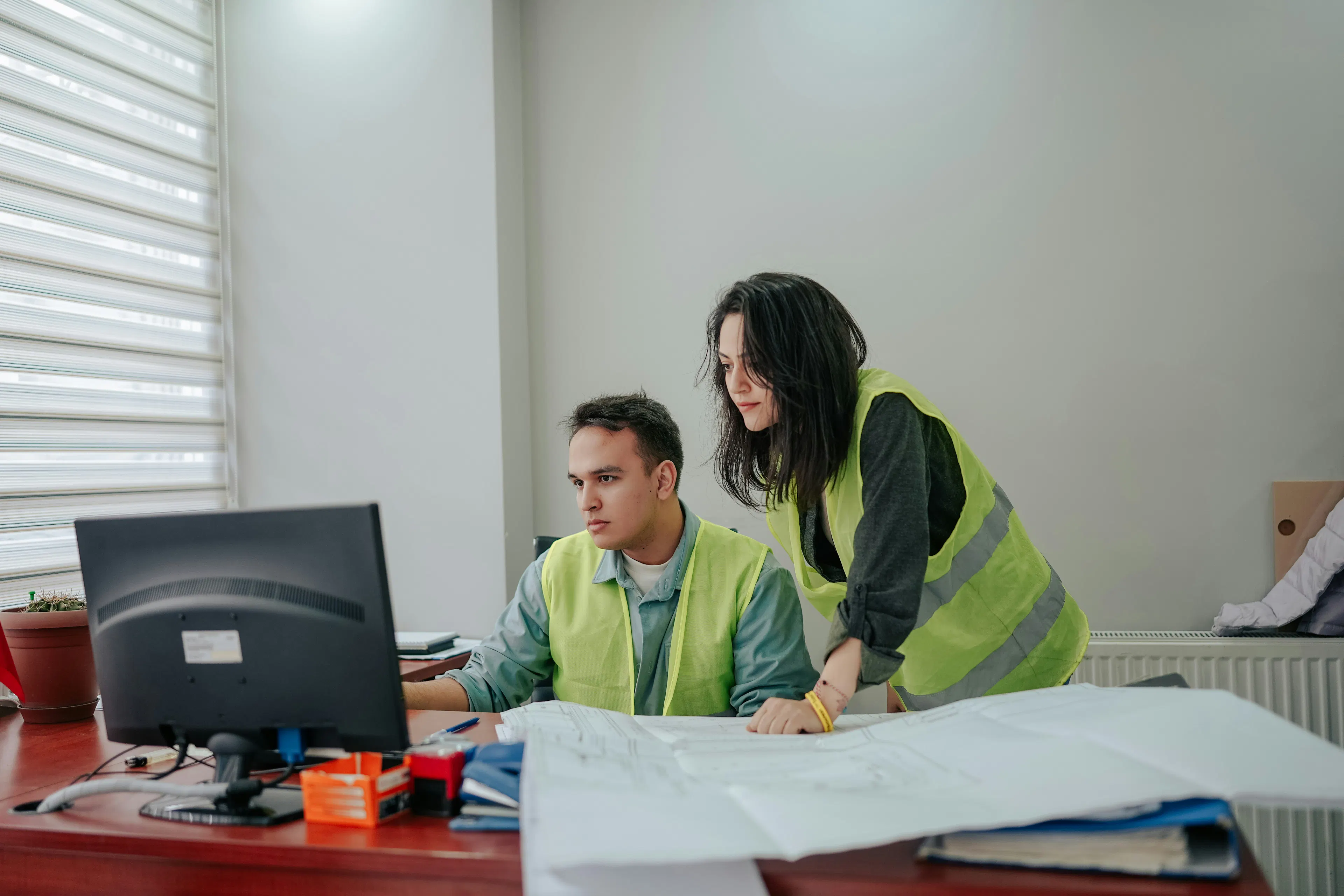 Two workers in hi-vis vests reviewing plans at a computer.