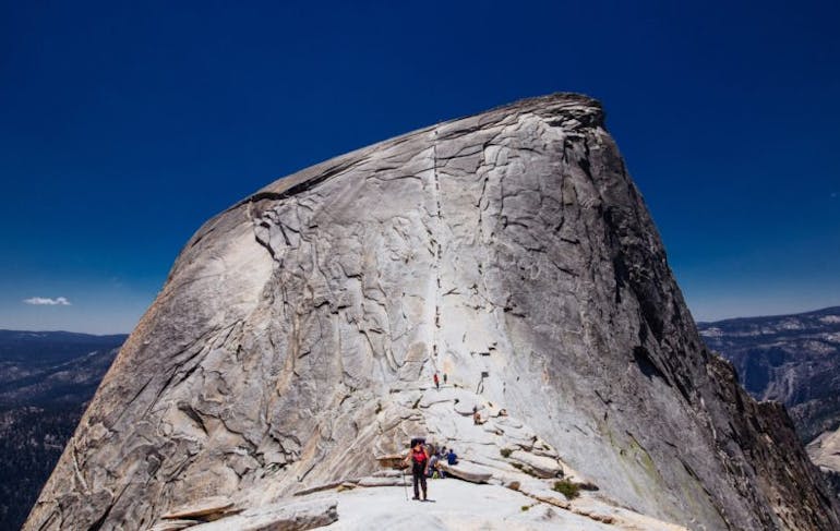 A giant grey boulder sits against a dark blue, cloudless sky. A person who looks small in scale stands in front of it.