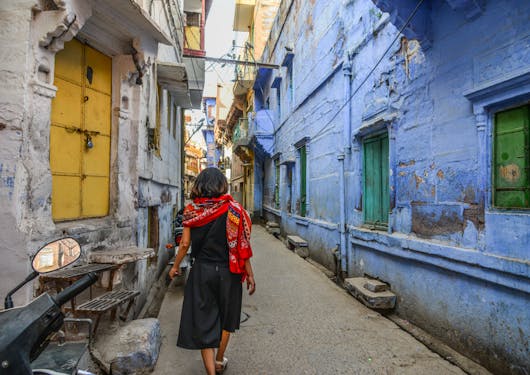 An adult women walks away from the camera through the streets of Old Jaipur on the Golden Triangle Cultural Tour of India. There is a yellow window to her left and blue buildings to the right. She is wearing a black short sleeve top and a black skirt with white sandals an a red scarf/wrap.