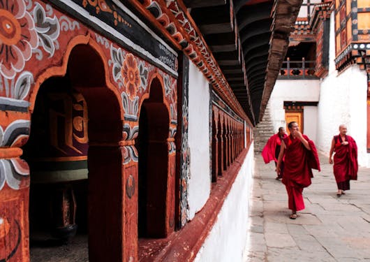 The inside of a brightly colored red and orange monastery is shown in Bhutan near Paro, with three monks in authentic and traditional red garbs and robes walk toward the camera on the right hand side.