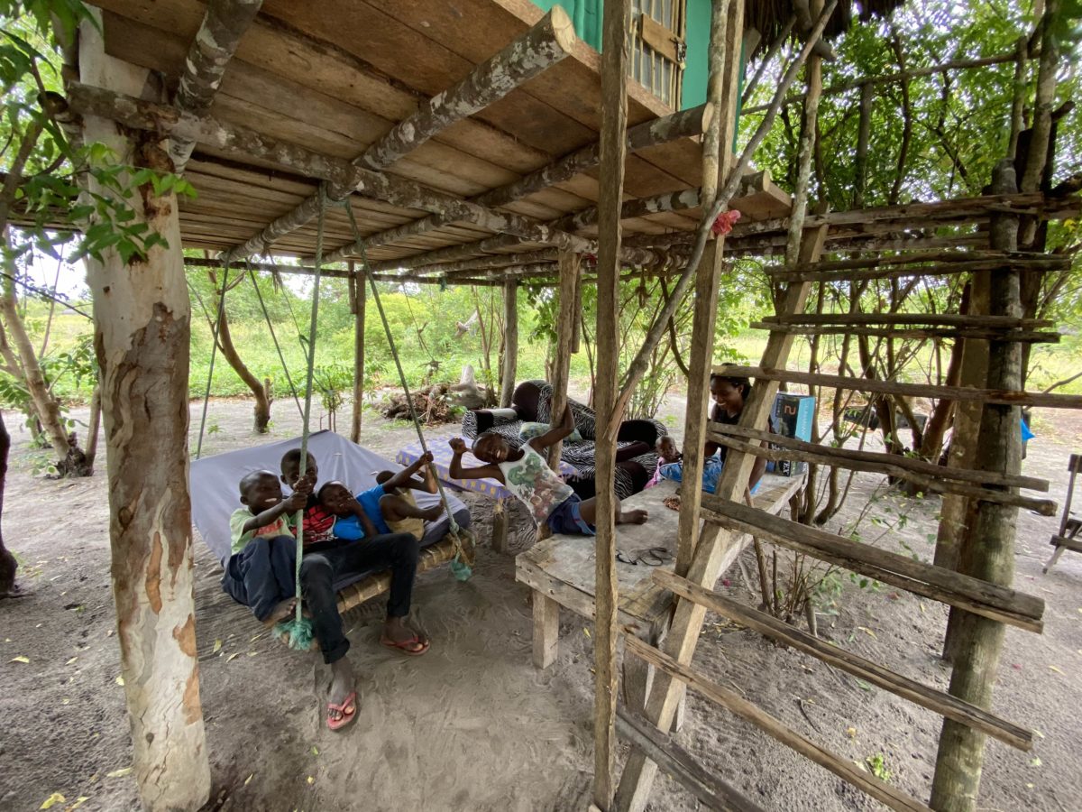 A group of 5 local Kenyan children sit under a stilted structure on a swing chair, smiling and enjoying themselves.