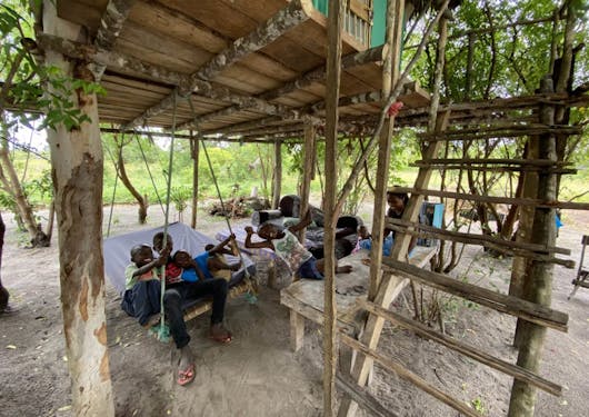 A group of 5 local Kenyan children sit under a stilted structure on a swing chair, smiling and enjoying themselves.