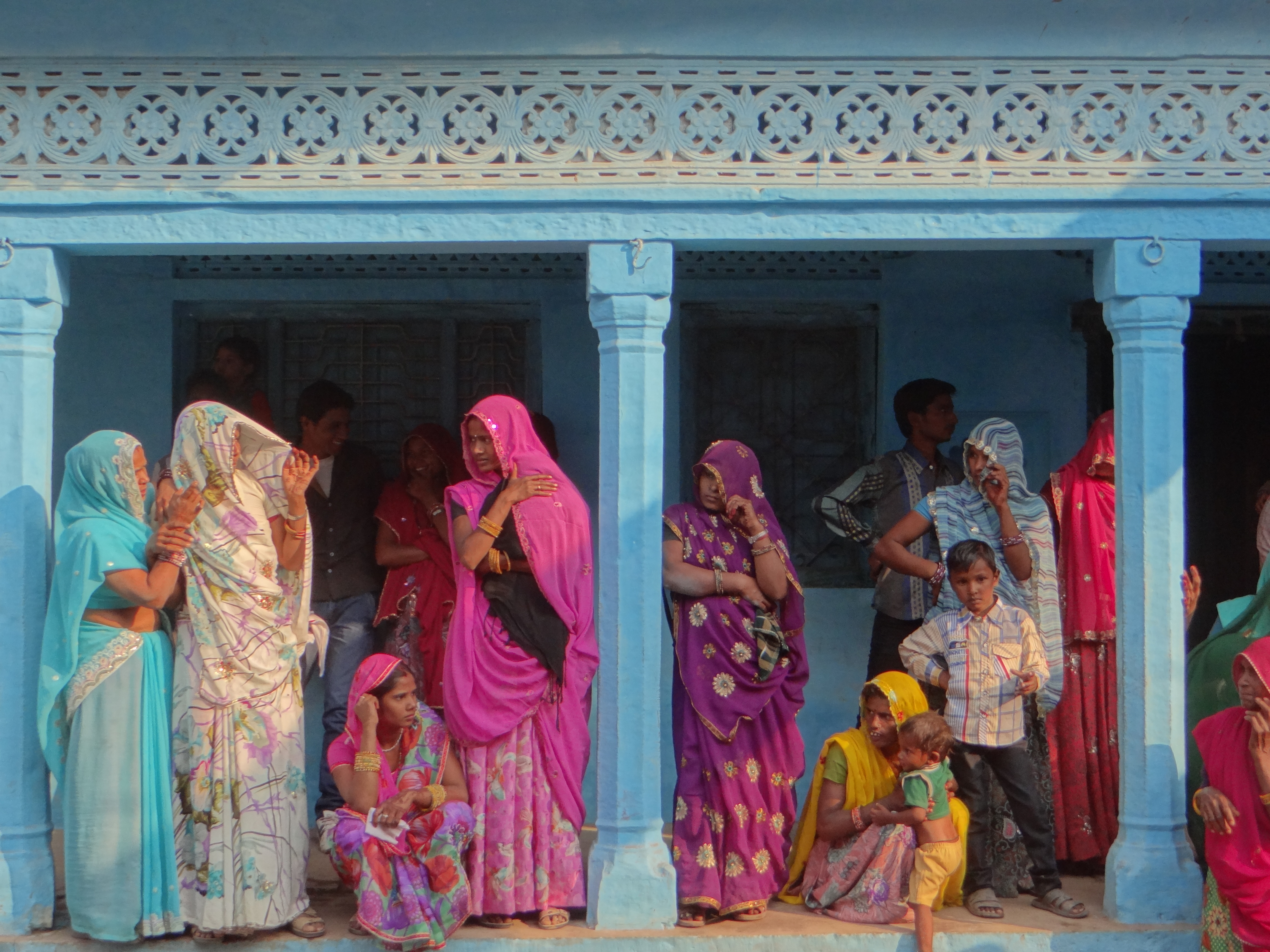 A group of mostly Indian women, with a young boy and a baby, stand in traditional colorful clothes of purples, pinks, and blues, under a blue awning supported by blue columns. 