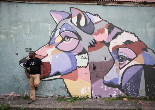 A street art mural of a mother and child wolf in light pastel colors including purples, pinks, and yellows, is shown against a light grey background. An adult black man wearing long khaki pants and a black t shirt is standing against the wall with one foot up against it propped behind him.