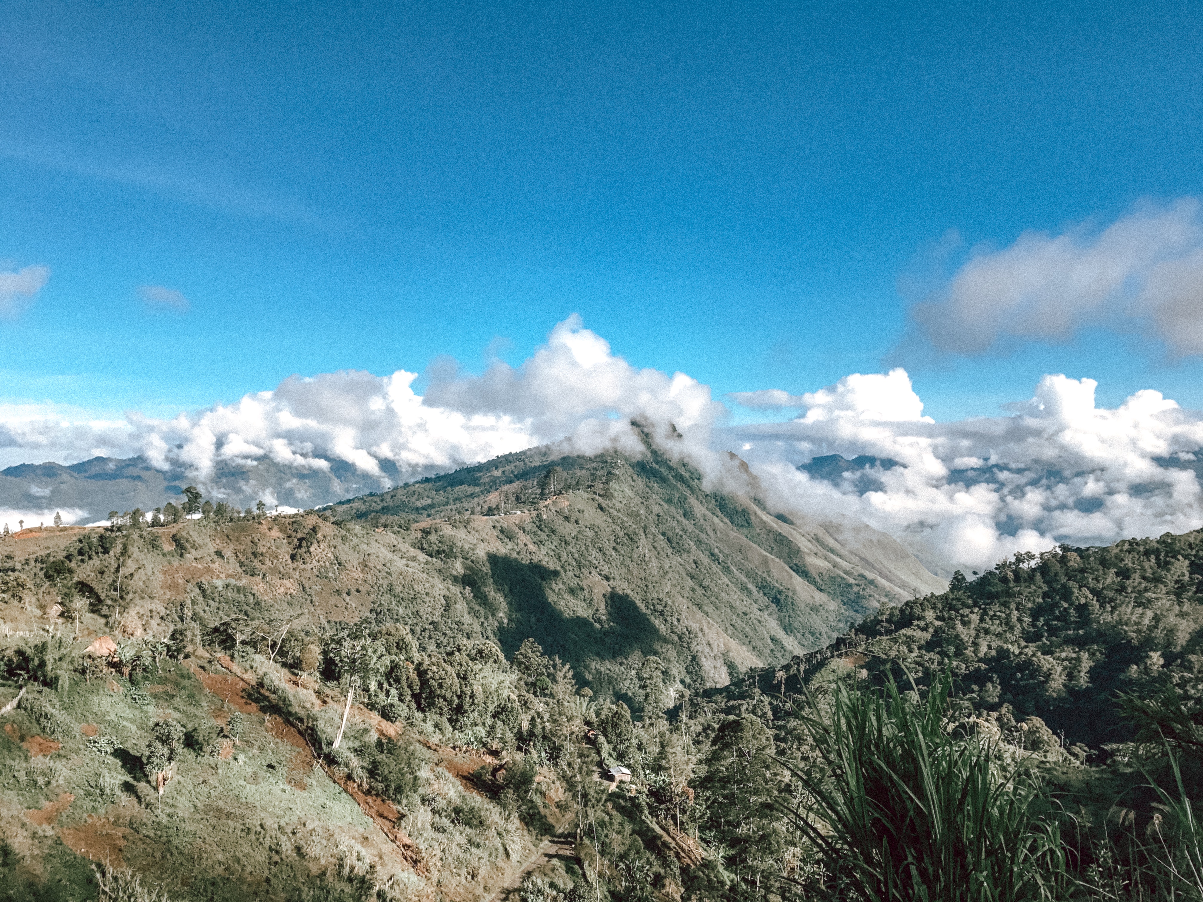 Papua New Guinea from up above - steep ascents of jungle greenery are shown as the peaks of mountains are in some fluffy white clouds with a blue sky above.