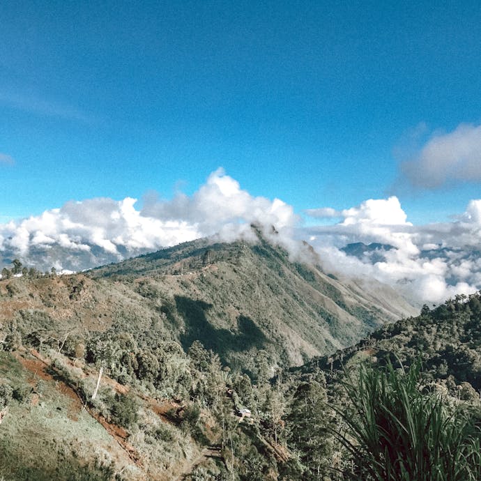 Papua New Guinea from up above - steep ascents of jungle greenery are shown as the peaks of mountains are in some fluffy white clouds with a blue sky above.
