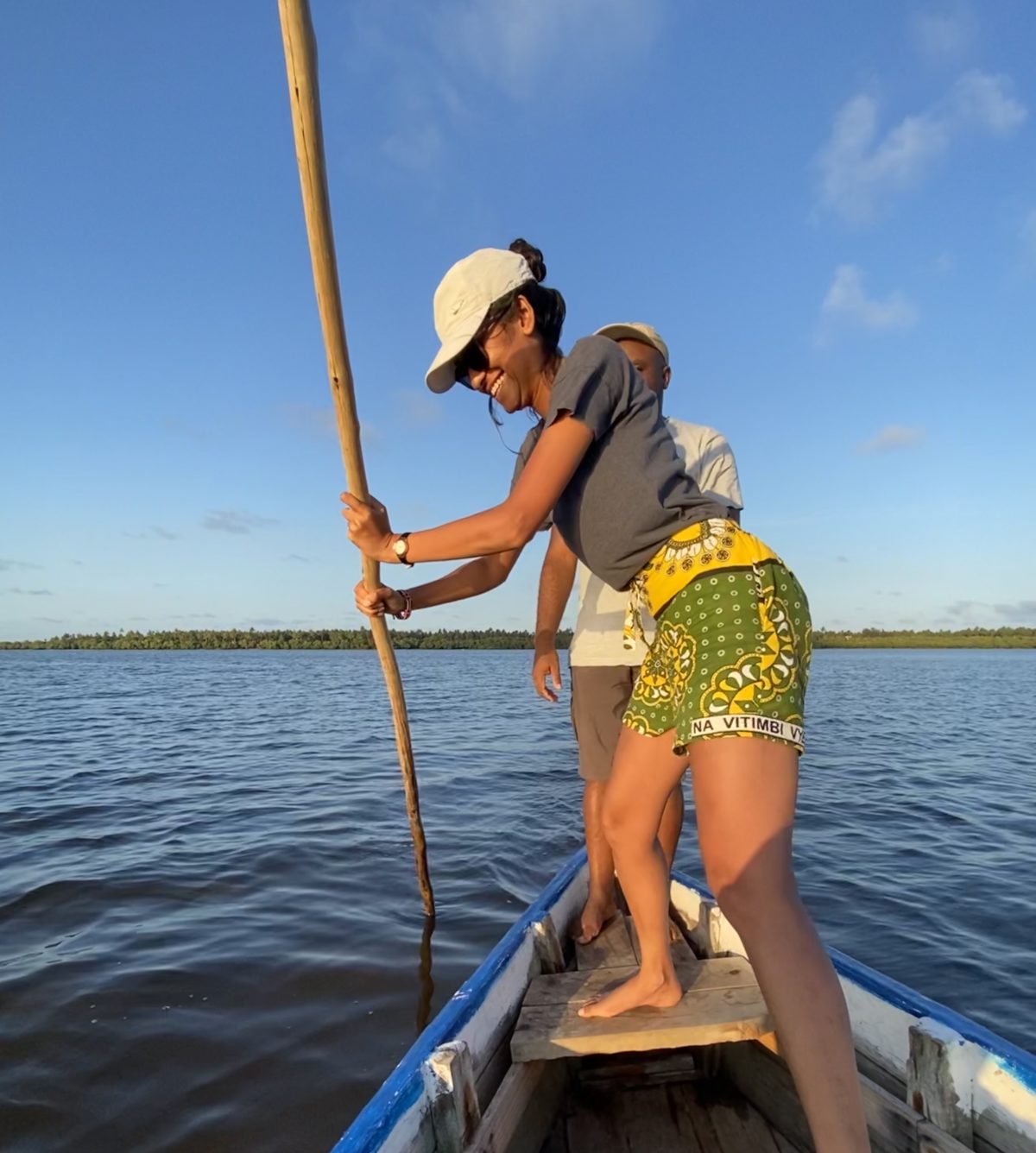 A local Kenyan woman uses a stick to push a small boat in the Mida Creek; she is wearing green and yellow shorts and a grey t shirt and a white cap that is shading her eyes as she grins widely. There is a man standing behind her observing. There is open water in the background with a land mass in the far back; the sky is blue with some light, wispy clouds.
