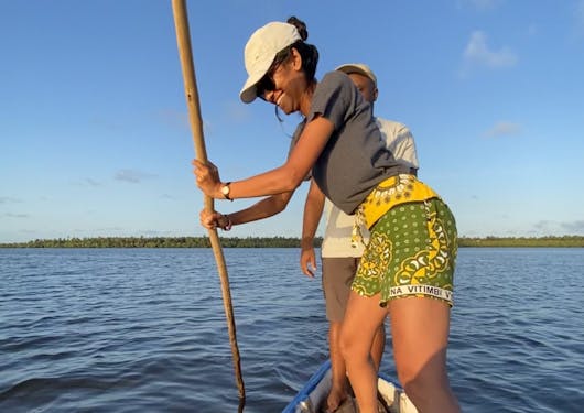 A local Kenyan woman uses a stick to push a small boat in the Mida Creek; she is wearing green and yellow shorts and a grey t shirt and a white cap that is shading her eyes as she grins widely. There is a man standing behind her observing. There is open water in the background with a land mass in the far back; the sky is blue with some light, wispy clouds.