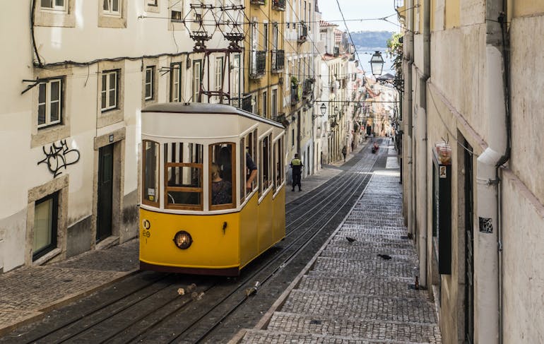 Yellow streetcar drives uphill surrounded by beige buildings in Lisbon, on a post guiding travelers about how to travel sustainably in Portugal.