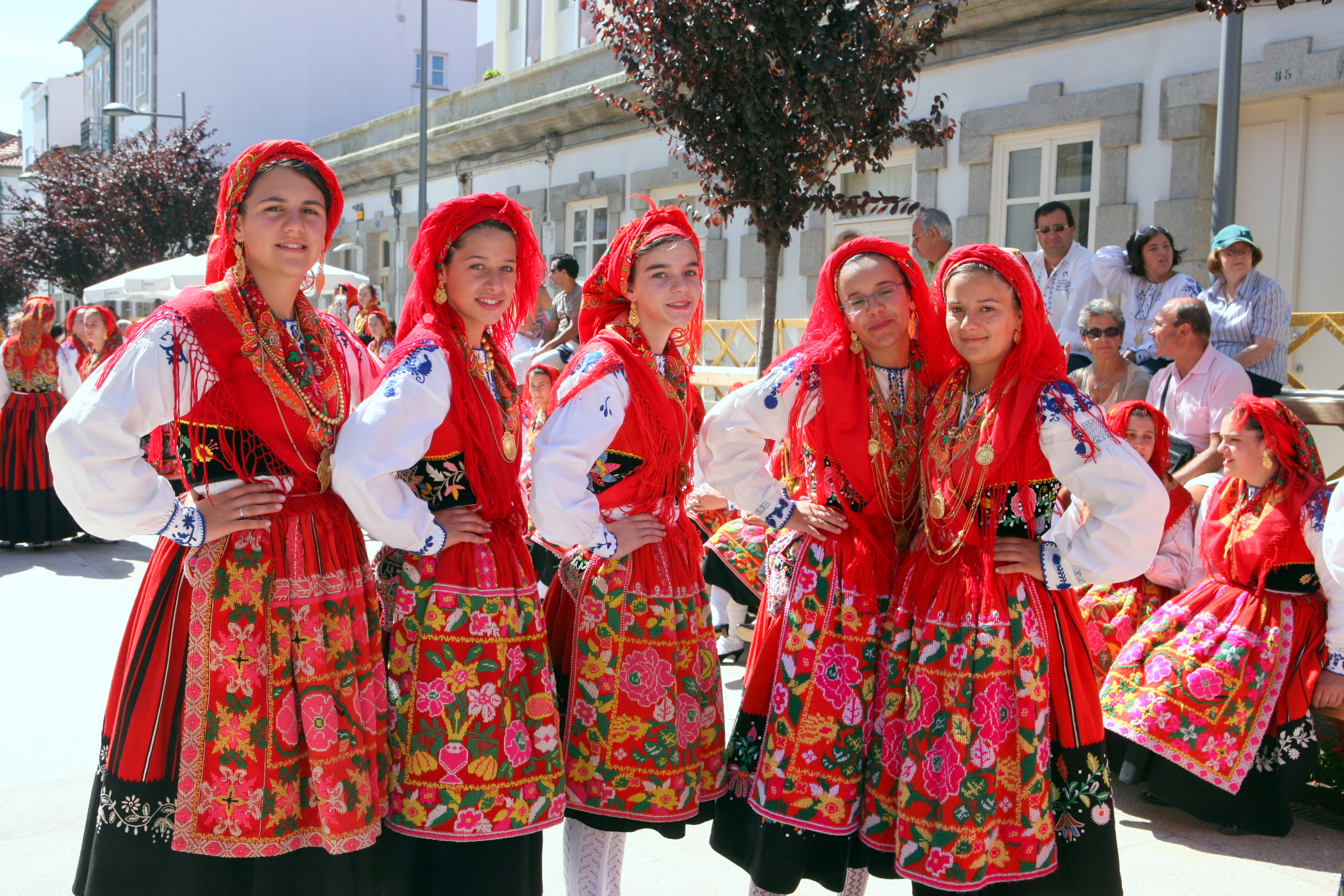 5 local Portuguese women dressed in traditional red and white (red dresses over long sleeve white blouses and red head wraps), stand in a row with their hands on their hips, smiling warmly.
