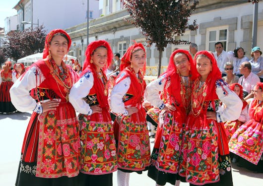 5 local Portuguese women dressed in traditional red and white (red dresses over long sleeve white blouses and red head wraps), stand in a row with their hands on their hips, smiling warmly.