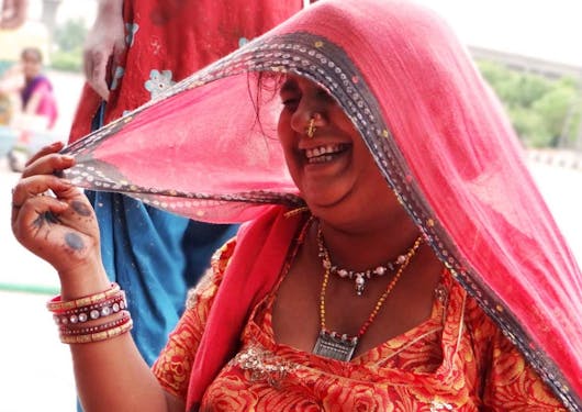An Indian women in traditional pink garb - a pink headcover and a reddish pink dress - wearing a gold nose ring, necklaces, and bracelets or bangles, smiles broadly while looking down and to the left side of the frame. Her face is partially covered by the head cover.
