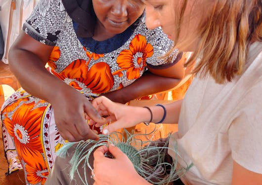 A local Black Keynan woman shows a White woman how to do some traditional crafts braiding threads.
