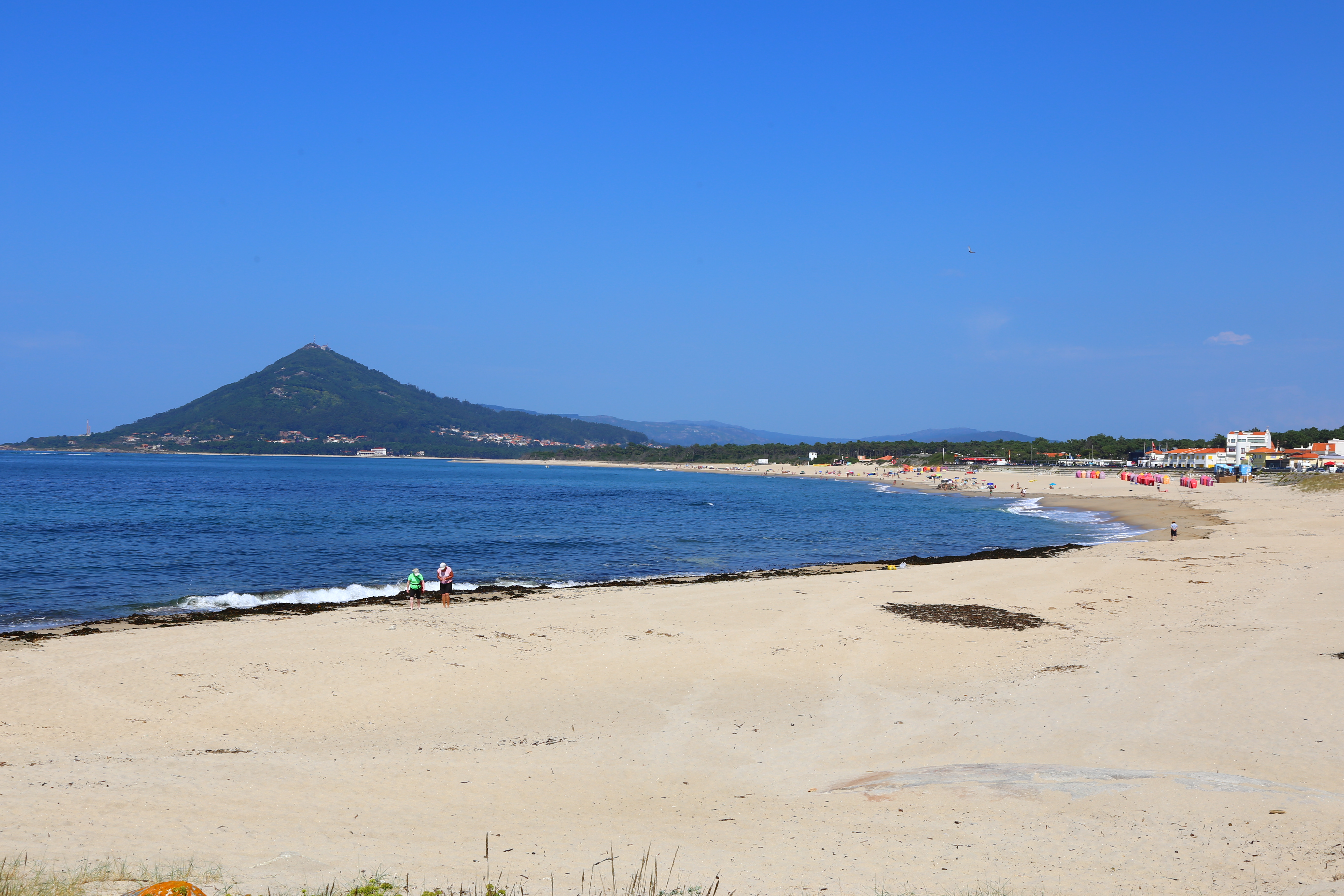 A flat, white sand beach meets the blue ocean water of the Atlantic in Portugal's Alto Minho region. A small, pointed hilltop is in the distance to the left. Colorful beach tents are off to the right, far away down the beach. It is sunny and the sky is blue and cloudless.
