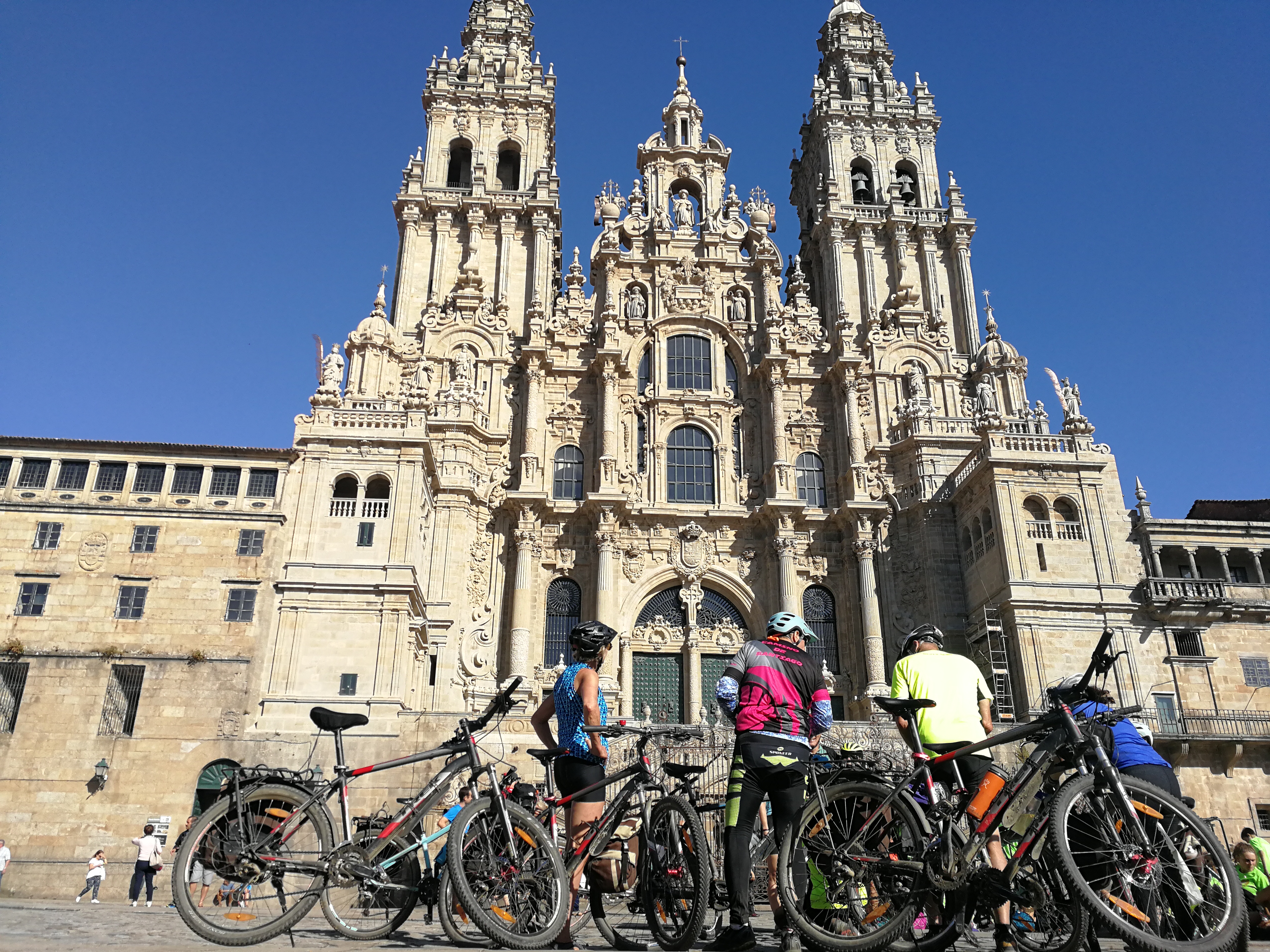 A group of 3 adult Caucasian bikers stand with their bikes in front of the majestic Cathedral de Santiago.
