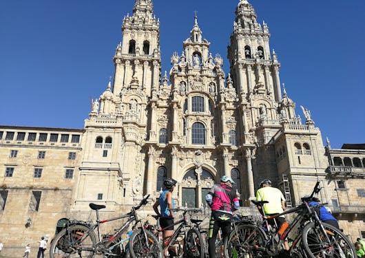 A group of 3 adult Caucasian bikers stand with their bikes in front of the majestic Cathedral de Santiago.