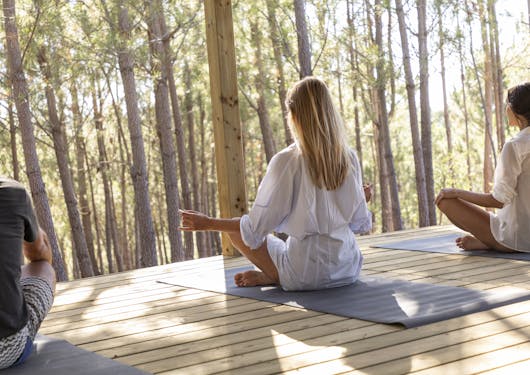 A Caucasian woman in a mid-length, white, long sleeve shirt dress sits in a meditative pose with her back to the camera. She sits on a yoga mat with a man sitting to her left and another Caucasian woman sitting to her right. They are on a slat wooden ground, overlooking thin trees with bright sunlight in the background.