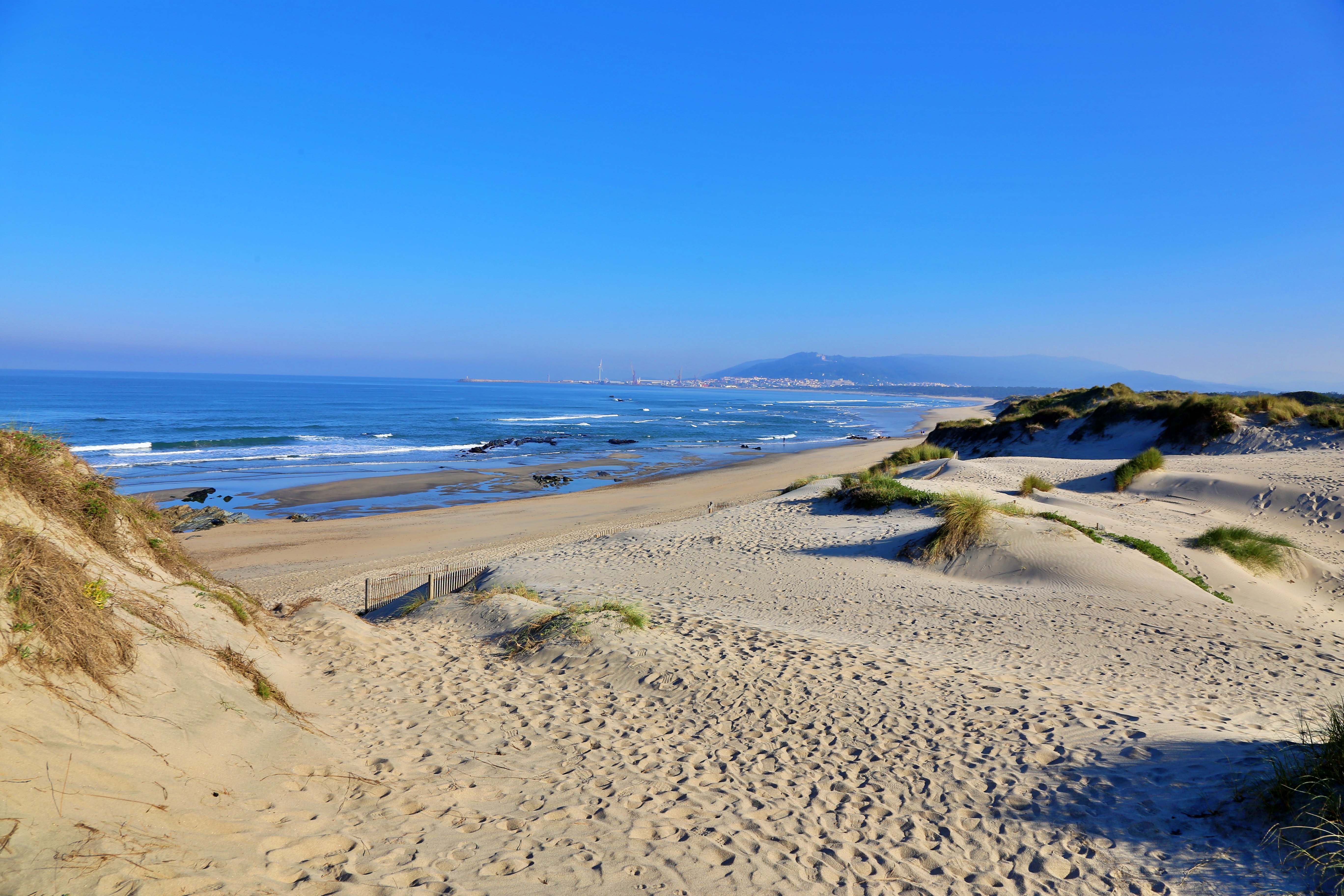 A white sand beach meets shallow, blue Atlantic ocean water under a blue sky in Portugal's Alto Minho region. The sky has no clouds and there are no people on the beach. The beach has some gentle sand mounds on it.