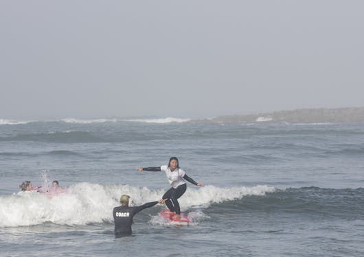 An adult Asian woman stands on a red surfboard and rides a wave while her instructor stands in nearby shallow water, giving coaching guidance. They are in the Atlantic Coast near southern Portugal at a sustainable surf retreat.
