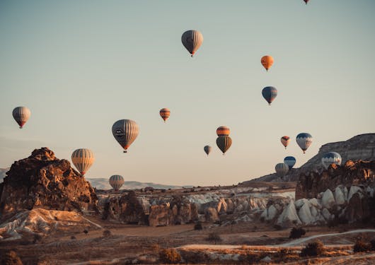 Hot air balloons float over the rocky landscape in Cappadocia on a cloudless morning. There is a warm light casting over the scene suggesting the sun is just starting to rise.