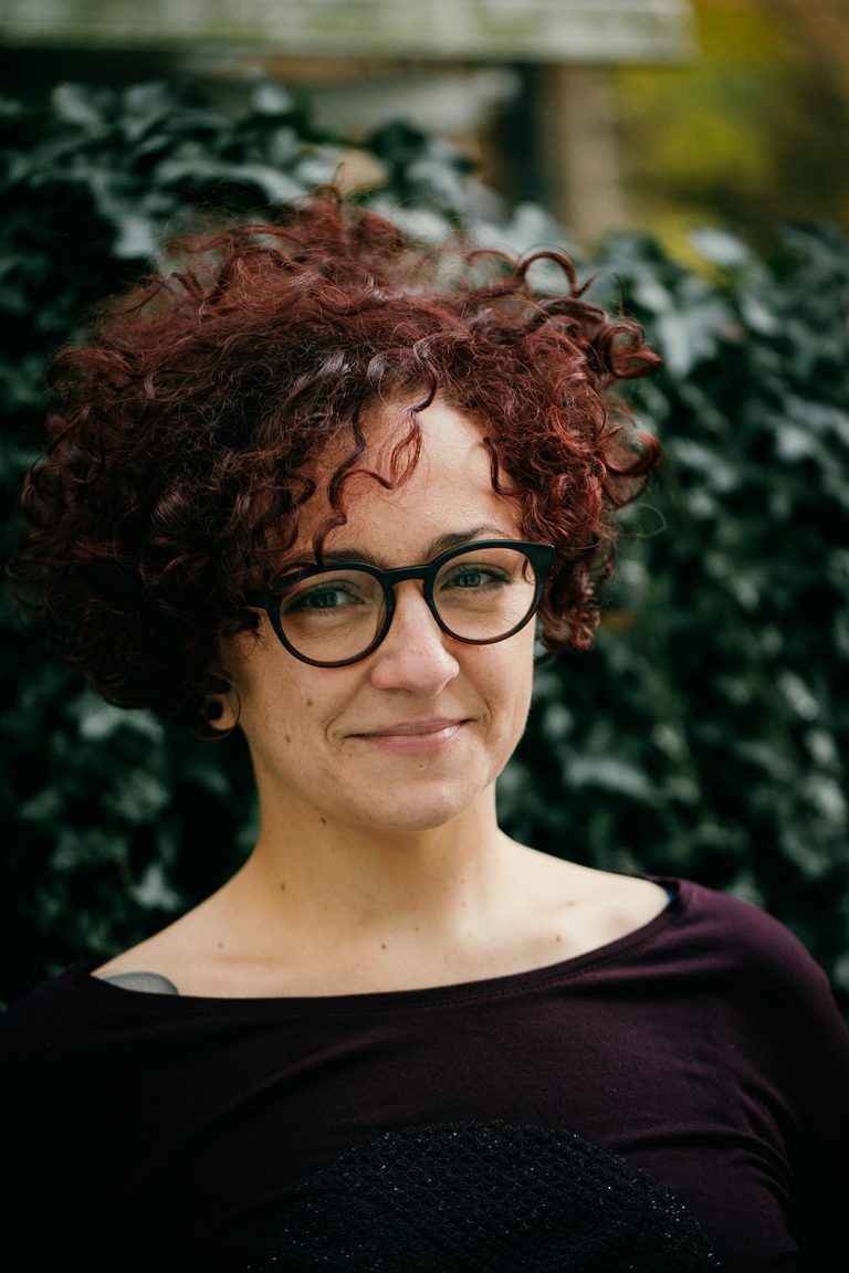 A headshot of a Caucasian woman with curly dark red hair and a dark top, wearing dark glasses and smiling with her mouth closes. She's standing in front of green foliage.