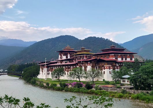 A monastery with white walls and a red roof in Punakha, Bhutan sits alongside a river surrounded by lunch greenery and rolling forested hills in the background.