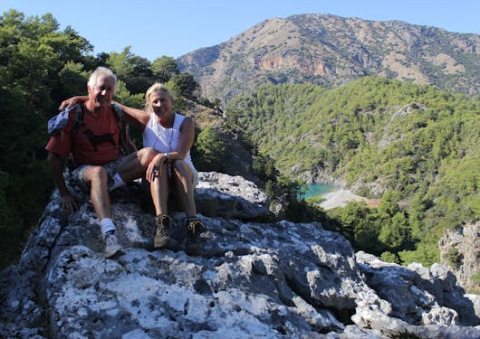 A caucasian couple, a man and woman, about in their 60's sit on top of a rock on top of a hill, with turquoise water down below past a hillside of green trees.