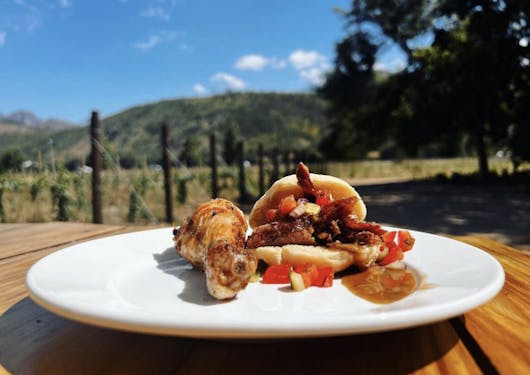 A closeup of a white plate with a cooked chicken drumstick on the left and a bread sandwich filled with traditional food sits on a wooden table, with green hills and trees blurry in the background.