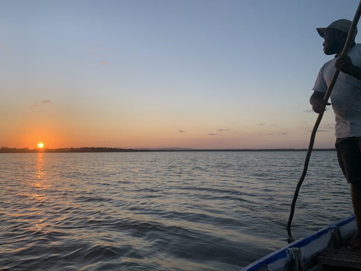 A beautiful orange sunset over Mida Creek in Kenya, as seen from a small boat on the water. There is a man on the right-hand side wearing a white t-shirt and a forward baseball hat using a stick to help steer and guide the boat.