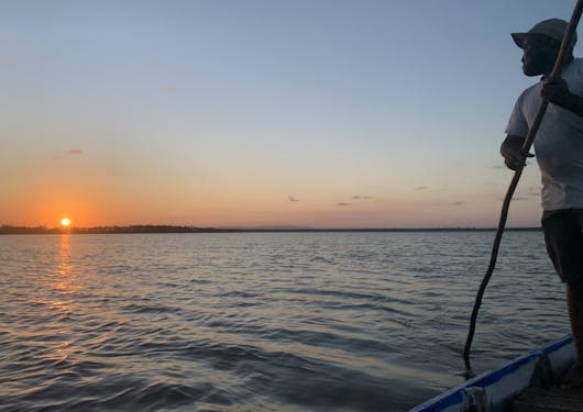 A beautiful orange sunset over Mida Creek in Kenya, as seen from a small boat on the water. There is a man on the right-hand side wearing a white t-shirt and a forward baseball hat using a stick to help steer and guide the boat.