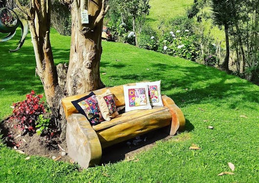 A wooden bench with 4 colorful, traditional pillows on it sits in front of a tree on a grassy lawn with more grass and greenery in the background. This is in the garden of the Villa Doris Country Lodge, a sustainable, eco-friendly lodge in Ecuador.